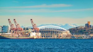 A scenic view of Seattle's waterfront featuring a stadium, cranes, and mountains.